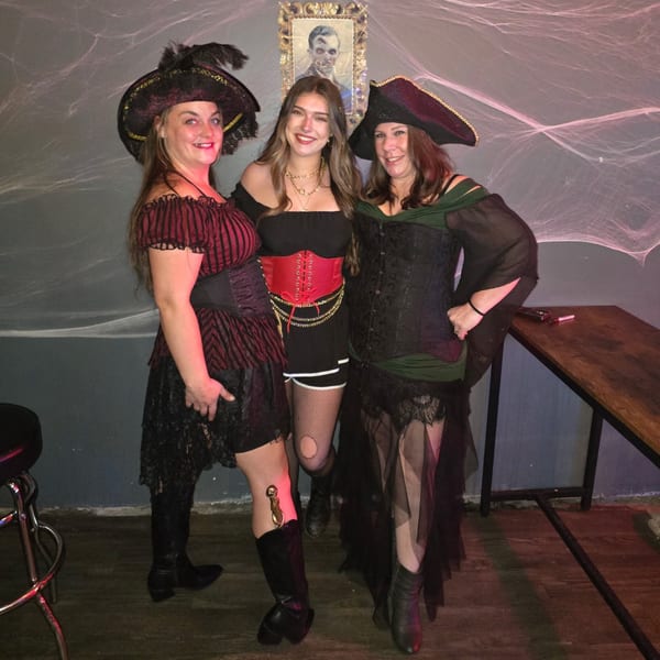 Three women in pirate costumes pose at Rendezvous Bar & Grill, with a spooky portrait and cobwebs.