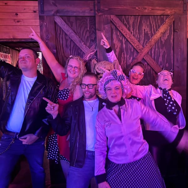 Six people dressed in 1950s-style outfits, including leather jackets and pink ladies jackets, posing and smiling.