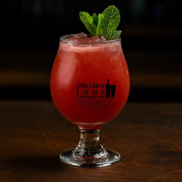A red cocktail with ice and mint garnish in a branded glass on a dark wooden table.