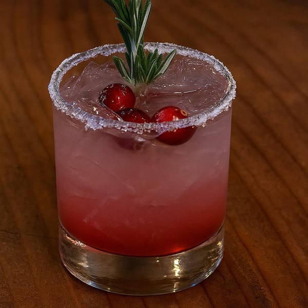 A cranberry cocktail with a sugared rim, garnished with fresh cranberries and a rosemary sprig.