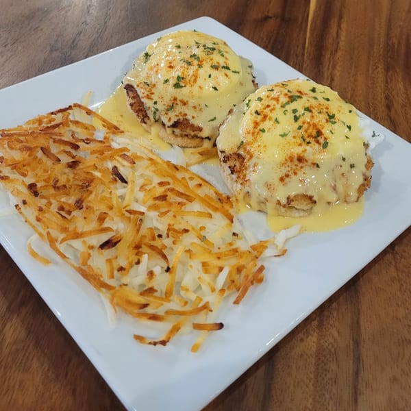 Two crab cake eggs Benedict with hollandaise sauce and hash browns on a white plate.