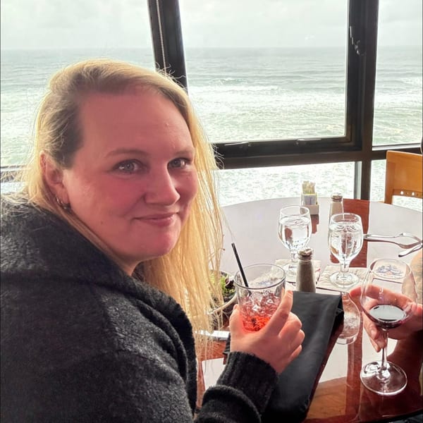 A woman with blonde hair smiles at the camera, holding a drink at a table overlooking the ocean.