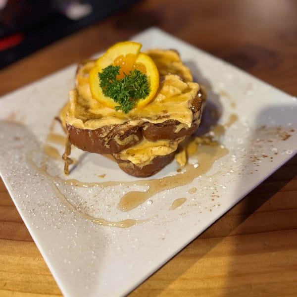 Two slices of French toast topped with orange cream, an orange slice, and parsley, drizzled with syrup.